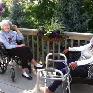 Two elderly individuals enjoy a sunny day on a porch, surrounded by greenery.