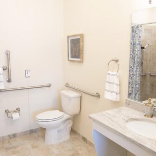 An accessible bathroom featuring a toilet with handrails, a sink with a mirror above, and a towel rack.