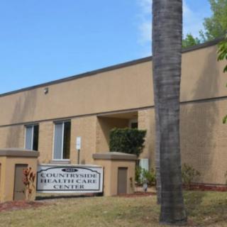 A health care building with a sign in front, surrounded by greenery and viewed on a clear day.