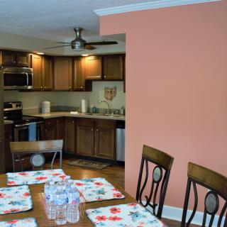 A cozy dining area with a wooden table and chairs, set with floral placemats and bottled water, adjacent to a kitchen featuring wooden cabinets and modern appliances.