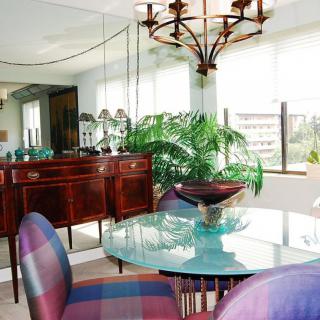 A well-lit dining room with a glass table and elegant chairs, featuring a large mirror and decorative plants.