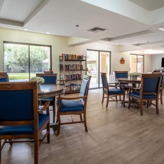 A cozy reading area with tables, chairs, and bookshelves illuminated by natural light through large windows.