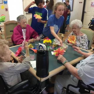 A group of elderly women engage in a crafts session with younger volunteers at a nursing home.