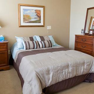A cozy bedroom featuring a neatly made bed with pillows and a comforter, accompanied by a nightstand with a lamp, a dresser with a mirror, and a landscape painting on the wall.