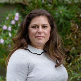 A woman standing in an outdoor setting with greenery in the background.