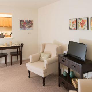 A cozy living room with an armchair, a TV on a dark wood stand, and a small dining area in the background.