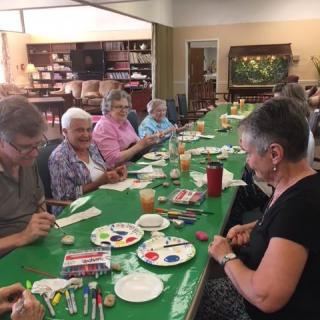 A group of seniors participates in a painting and craft activity around a large table.