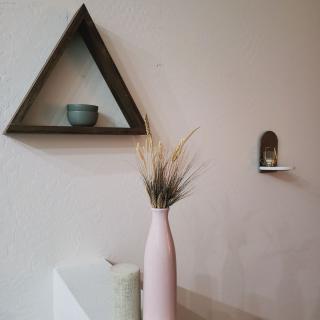A minimalist interior featuring a wooden triangle shelf with a ceramic bowl, alongside a pink vase filled with dry grass.