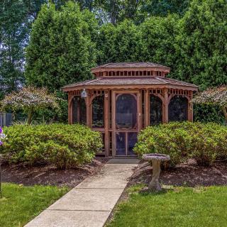 A charming wooden gazebo surrounded by lush greenery and bushes, with a stone pathway leading to it.