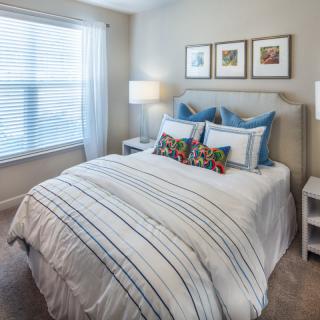 A cozy and well-decorated bedroom with a neatly made bed, featuring striped bedding and colorful pillows. The room is brightened by natural light from a large window and complemented by framed artwork above the headboard.