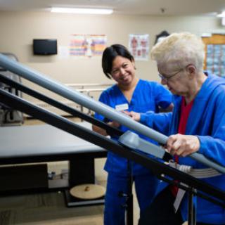 A nurse assists an elderly patient during a physical therapy session, providing support as the patient uses a handrail.