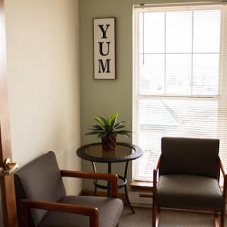 A cozy waiting area featuring two chairs, a small table with a plant, and natural light streaming through the window.