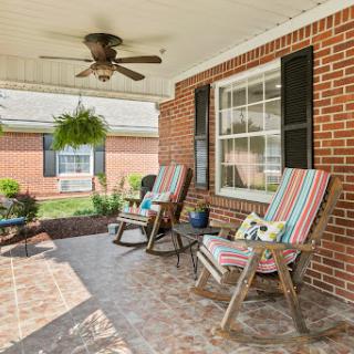 A cozy porch featuring colorful rocking chairs, a ceiling fan, and potted plants, perfect for enjoying a sunny day.