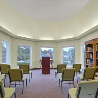 A small meeting room with a podium and several chairs arranged in rows, surrounded by windows and a bookshelf.