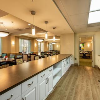 A spacious dining room with a long counter, tables, and chairs, featuring warm lighting and wood flooring.