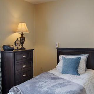 A cozy bedroom with a single bed, featuring a blue pillow and a dresser topped with a lamp.