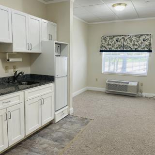A small apartment interior featuring a kitchen with white cabinets, a sink, and a refrigerator. The room has carpet flooring and a window allowing natural light.