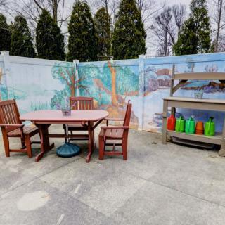 An outdoor patio with a wooden table and chairs, adjacent to a barbecue grill, set against a mural-painted fence.