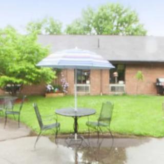 A patio setup with a table and chairs under an umbrella, situated in a garden area with wet ground.