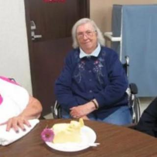 An elderly woman sitting in a wheelchair at a table with a slice of cake, possibly celebrating an occasion.