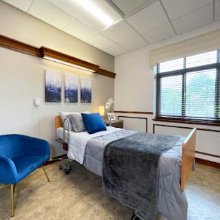 A well-lit hospital room featuring a bed with gray bedding, a blue accent chair, and artwork on the walls.