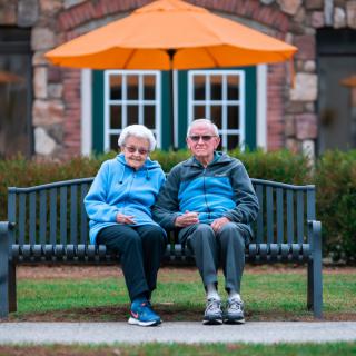 An elderly couple sitting together on a park bench under an orange umbrella.