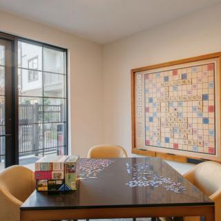 A cozy room with a table set up for assembling a puzzle, surrounded by chairs and artwork on the wall, illuminated by natural daylight.