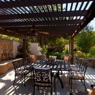 A cozy outdoor patio with a pergola providing shade over table and chairs, surrounded by lush garden greenery.