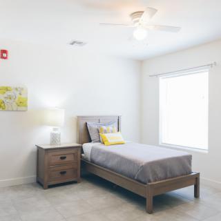 A bright and minimalist bedroom featuring a single bed with a nightstand and lamp, illuminated by natural light from a large window.