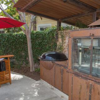 An outdoor kitchen setup featuring a barbecue grill, wooden cabinetry, and a vibrant red umbrella shaded area.
