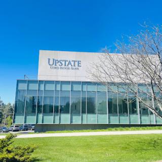 A modern glass-fronted building with the Upstate logo, surrounded by greenery and a clear blue sky.
