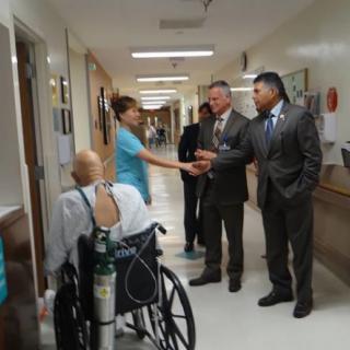 A nurse greets visitors in a hospital corridor as a patient in a wheelchair looks on.