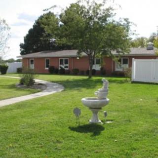 A suburban house with a large lawn featuring a birdbath and pathway, surrounded by trees and a white fence.
