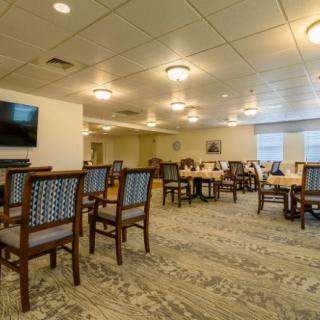 A spacious dining room with tables and chairs, well-lit with overhead lights, featuring a carpeted floor and a television on the wall.