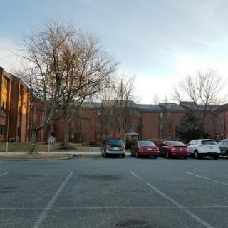 A view of a brick building beside a parking lot with several cars parked and bare trees in the foreground.