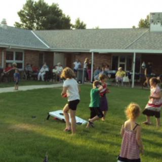 Children playing outdoors at a community gathering on a grassy area in front of a building.