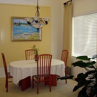 A cozy dining room with a table covered by a white cloth, surrounded by wooden chairs. A painting hangs on the wall, and a potted plant is placed near the window.