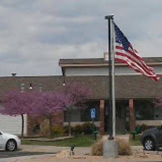 A view of a residential building showcasing an American flag and vibrant purple trees against a cloudy backdrop.