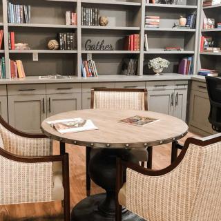 A cozy reading area featuring a round table surrounded by chairs, set against bookshelves filled with various items.