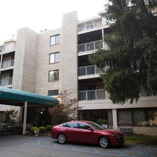 A multi-story apartment building with a red car parked in front and trees surrounding the entrance.