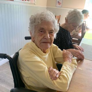 An elderly woman sits in a wheelchair, smiling warmly in a care facility, enjoying the company of others.