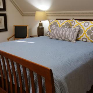 A cozy attic bedroom with a neatly made bed, decorative pillows, and a warm lamp.