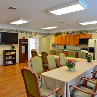 A spacious dining room with a table and chairs, adjacent to a kitchen area, decorated with flowers and featuring a television and bookshelf.