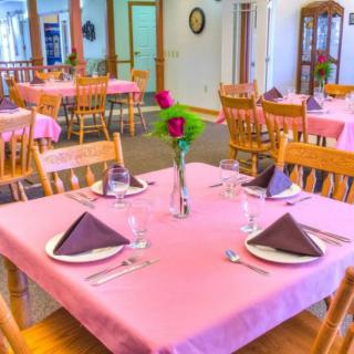 A cozy restaurant dining area with tables set for guests, featuring pink tablecloths and roses in vases.