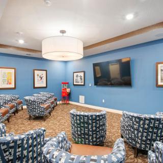 A cozy lounge area with patterned chairs arranged around a large television on blue-painted walls, adorned with framed artwork and illuminated by a central overhead light fixture.