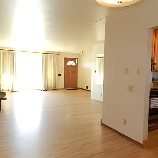 A bright living room with hardwood flooring, natural light streaming through large windows, and an entrance to the kitchen.