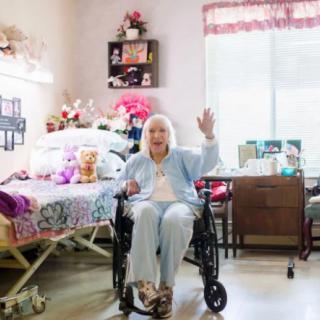 An elderly woman in a wheelchair waves in a cozy nursing home room, surrounded by flowers and personal items.