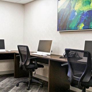 A modern office workspace featuring three desktop computers, ergonomic chairs, and a colorful abstract painting on the wall.