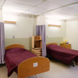 A hospital room with two twin beds, separated by a curtain, and simple wooden furniture.