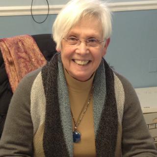 A smiling person with white hair and glasses, wearing a scarf indoors.